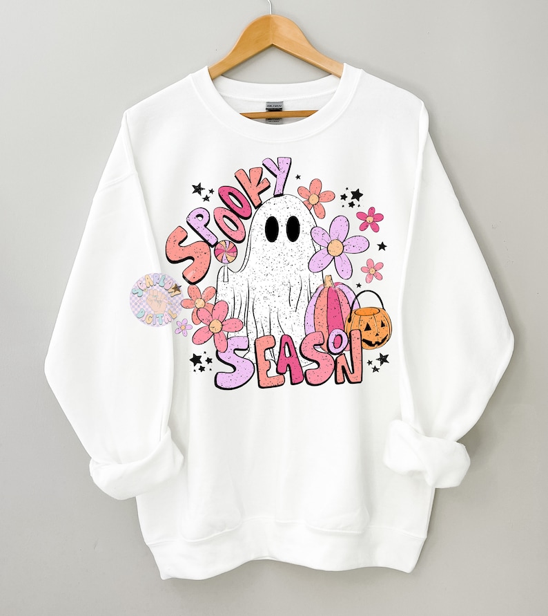 Pu&ograve; includere: Felpa bianca con un design grafico rosa, arancione e bianco di un fantasma, fiori, zucche e il testo "Spooky Season".