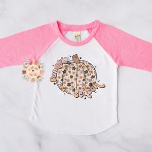 Pu&ograve; includere: Una maglietta da baseball bianca e rosa con un design a zucca floreale e il testo "Pumpkin Season".