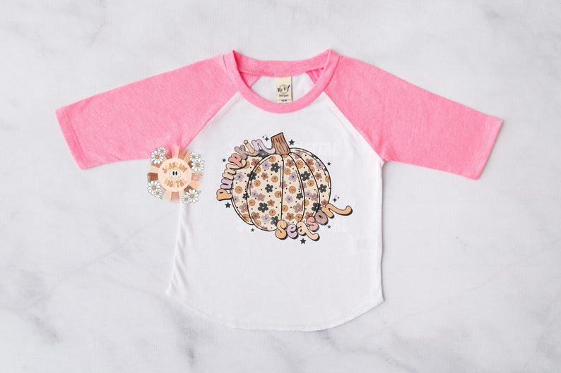 Pu&ograve; includere: Una maglietta da baseball bianca e rosa con un design a zucca floreale e il testo "Pumpkin Season".
