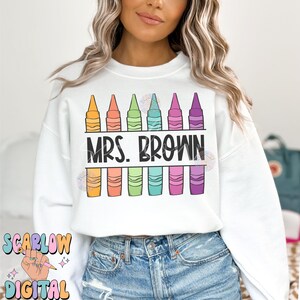 以下が含まれることがあります： 白いスウェットシャツに、カラフルなクレヨンの列と黒い文字で「MRS. BROWN」のデザインが施されています。