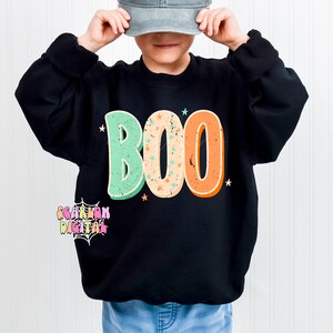 Peut inclure: Un enfant portant un sweat-shirt noir avec le mot "BOO" en lettres r&eacute;tro color&eacute;es et des &eacute;toiles. L'enfant porte une casquette de baseball grise et un jean bleu.