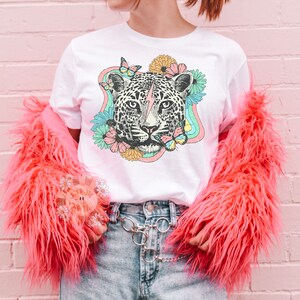May include: White t-shirt with a graphic of a leopard's face with a lightning bolt, surrounded by flowers and butterflies in a rainbow-colored design.