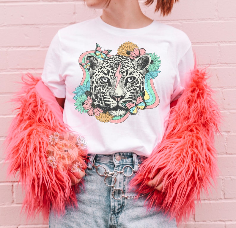 May include: White t-shirt with a graphic of a leopard's face with a lightning bolt, surrounded by flowers and butterflies in a rainbow-colored design.