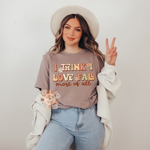 Op de afbeelding: Een vrouw draagt een grijs T-shirt met de tekst "I think I love *fall* most of all" in kleurrijke letters. Ze draagt een witte hoed en een witte cardigan.