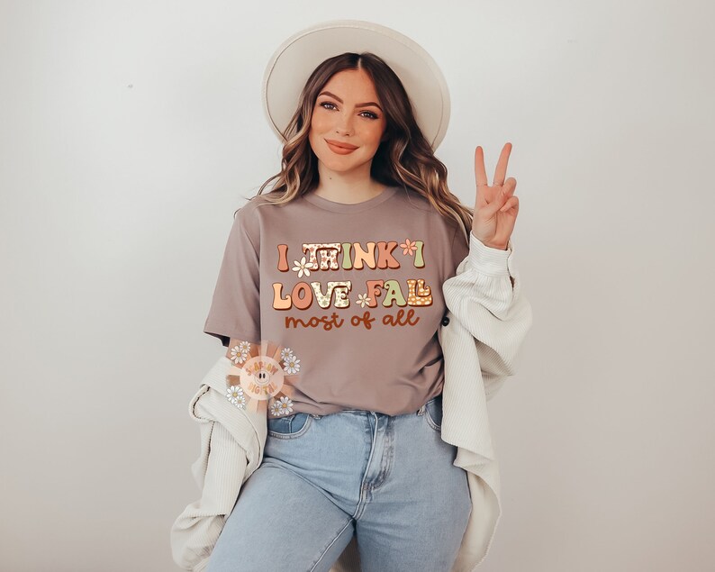 Op de afbeelding: Een vrouw draagt een grijs T-shirt met de tekst "I think I love *fall* most of all" in kleurrijke letters. Ze draagt een witte hoed en een witte cardigan.