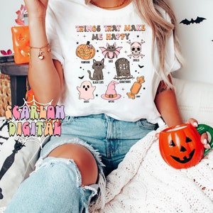 Puede incluir: Camiseta blanca con un dise&ntilde;o gr&aacute;fico de iconos de Halloween y el texto "Things That Make Me Happy". Los iconos incluyen calabazas, murci&eacute;lagos, calaveras, gatos negros, fantasmas, brujas, cementerios y caramelos.