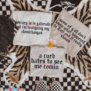 May include: Three t-shirts with text are displayed on a patterned surface. One shirt is white with the text "mercury is in gatorade and I'm realigning my chimichangas." Another is tan with the text "some of y'all don't know what happened to Earl and it really shows." The third is gray with the text "a curb hates to see me comin."
