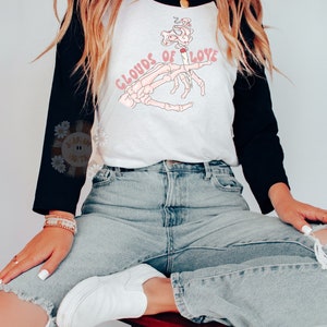 May include: A white and black raglan style shirt with the text "CLOUDS OF LOVE" and a graphic of a skeleton hand holding a lit cigarette. The shirt is paired with light wash jeans and white shoes.