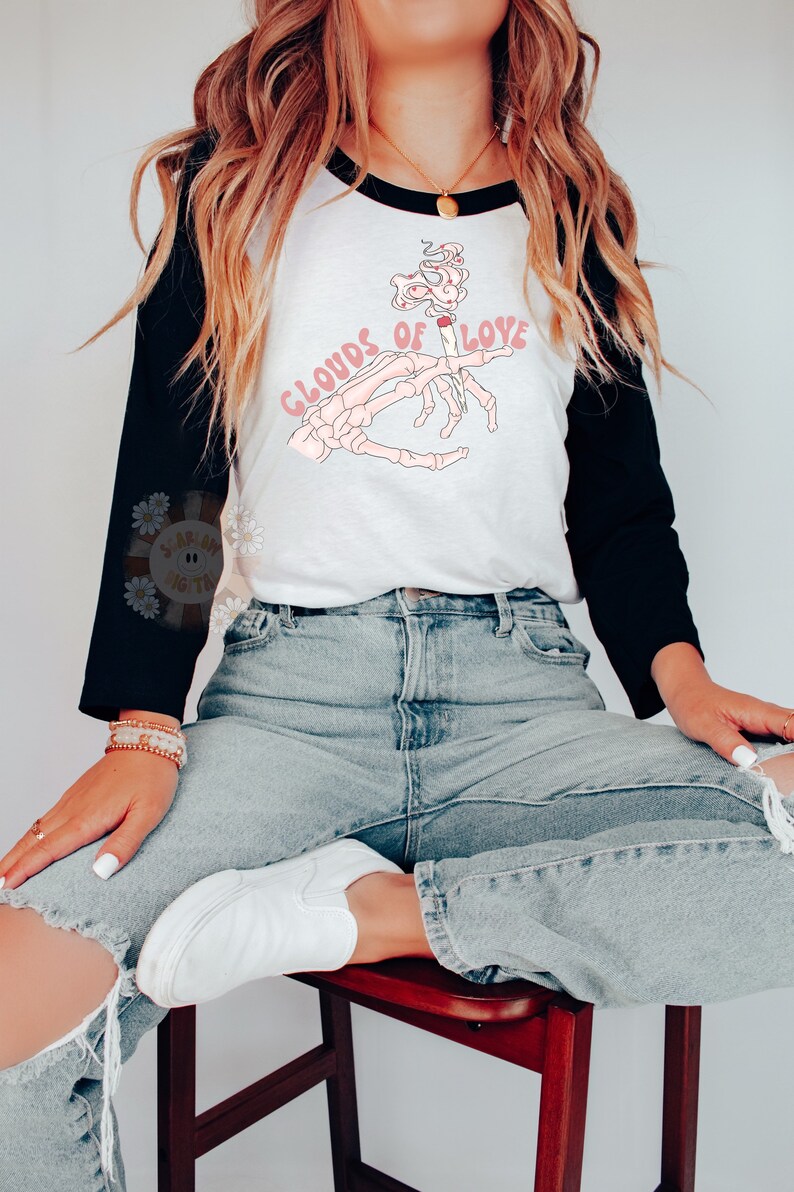 May include: A white and black raglan style shirt with the text "CLOUDS OF LOVE" and a graphic of a skeleton hand holding a lit cigarette. The shirt is paired with light wash jeans and white shoes.
