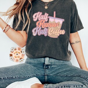 May include: Dark gray t-shirt with the text "Half Human Half Coffee" in pink, orange, and purple. The graphic includes a coffee cup with a pink lid and a straw. The shirt is paired with blue jeans and white sneakers.