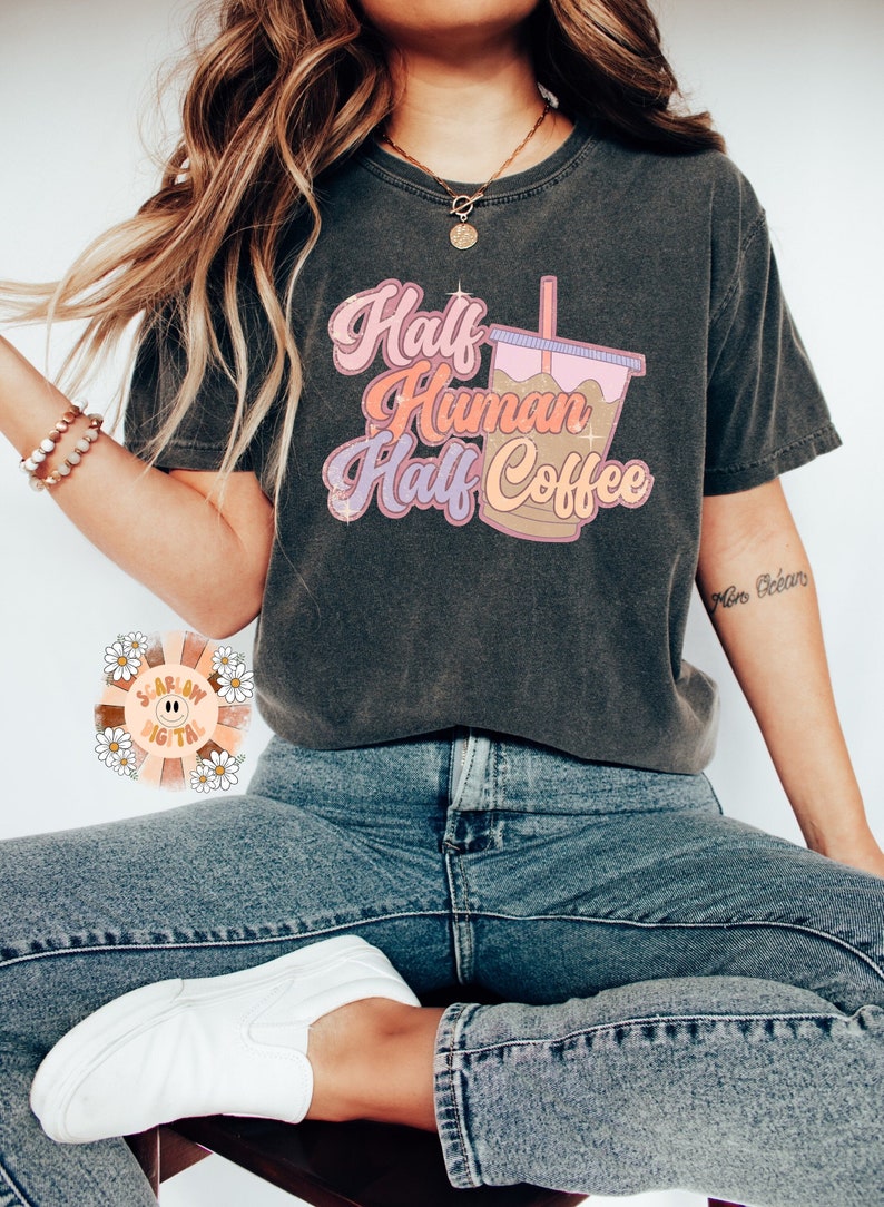 May include: Dark gray t-shirt with the text "Half Human Half Coffee" in pink, orange, and purple. The graphic includes a coffee cup with a pink lid and a straw. The shirt is paired with blue jeans and white sneakers.