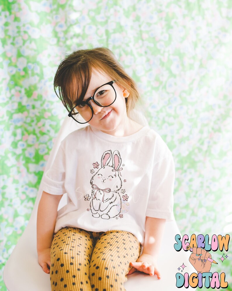 May include: A young child wearing a white t-shirt with a pink and white bunny graphic. The child is wearing brown and white polka dot pants and black glasses. The child is sitting on a white chair. The background is a green floral pattern.