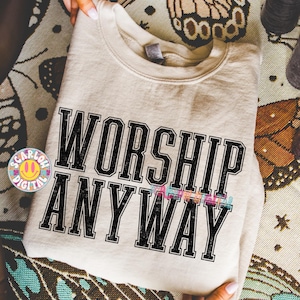 May include: A beige sweatshirt with the words "WORSHIP ANYWAY" in large, black, block letters. The word "ANYWAY" has a colorful, striped design. The sweatshirt is folded and held by hands.