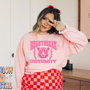 Pu&ograve; includere: Felpa rosa con una grafica di un leopardo rosa con occhi a forma di cuore e il testo "Heartbreak University".