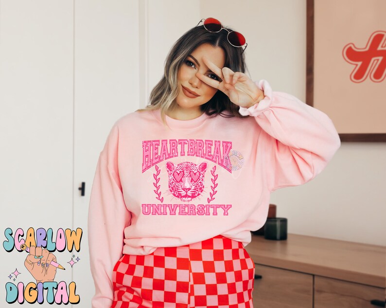 Pu&ograve; includere: Felpa rosa con una grafica di un leopardo rosa con occhi a forma di cuore e il testo "Heartbreak University".