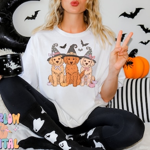 K&ouml;nnte beinhalten: Wei&szlig;es T-Shirt mit einem Aufdruck von drei Golden Retrievern, die Hexenh&uuml;te tragen und K&uuml;rbisse halten. Die Hunde tragen rosa und lila Schleifen. Der Aufdruck ist auf wei&szlig;em Hintergrund.