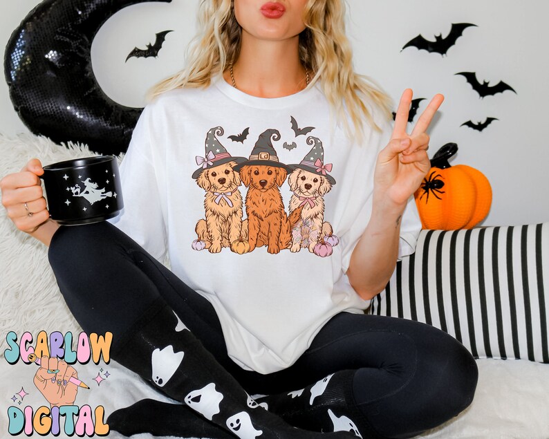K&ouml;nnte beinhalten: Wei&szlig;es T-Shirt mit einem Aufdruck von drei Golden Retrievern, die Hexenh&uuml;te tragen und K&uuml;rbisse halten. Die Hunde tragen rosa und lila Schleifen. Der Aufdruck ist auf wei&szlig;em Hintergrund.