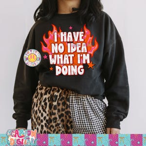 Puede incluir: Sudadera negra con el texto "I HAVE NO IDEA WHAT I'M DOING" en blanco y naranja, rodeado de llamas y estrellas. La sudadera se combina con pantalones con estampado de leopardo y cuadros. Un logotipo es visible.