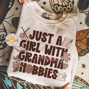 K&ouml;nnte beinhalten: Beiges Sweatshirt mit dem Text "JUST A GIRL WITH GRANDMA HOBBIES" in Braun. Das Design umfasst Illustrationen von Stricknadeln, einem Wollkn&auml;uel, einem Buch, einer Topfpflanze, einem Kuchen, einer N&auml;hmaschine und Puzzleteilen.