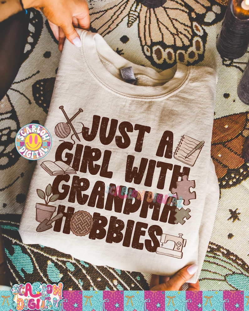 K&ouml;nnte beinhalten: Beiges Sweatshirt mit dem Text "JUST A GIRL WITH GRANDMA HOBBIES" in Braun. Das Design umfasst Illustrationen von Stricknadeln, einem Wollkn&auml;uel, einem Buch, einer Topfpflanze, einem Kuchen, einer N&auml;hmaschine und Puzzleteilen.