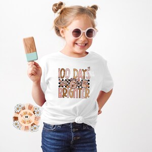 May include: A white t-shirt with a pink and brown graphic that says "100 Days Brighter". The graphic features a checkered pattern and a floral design.
