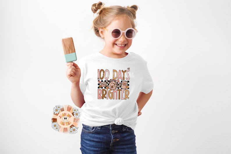 May include: A white t-shirt with a pink and brown graphic that says "100 Days Brighter". The graphic features a checkered pattern and a floral design.