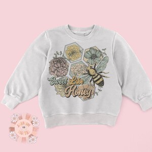 May include: A white sweatshirt with a graphic design of a bee and flowers in a honeycomb pattern. The text "Sweet Like Honey" is printed in a retro font.