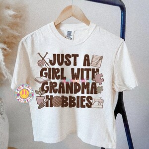 K&ouml;nnte beinhalten: Cremefarbenes T-Shirt mit dem Text "JUST A GIRL WITH GRANDMA HOBBIES" in Braun und bunten Buchstaben. Das Design zeigt Illustrationen von Stricknadeln, Notizbuch, Buch, Puzzleteil, Topfpflanze, N&auml;hmaschine und Garnkn&auml;uel, mit metrischen Ma&szlig;en.