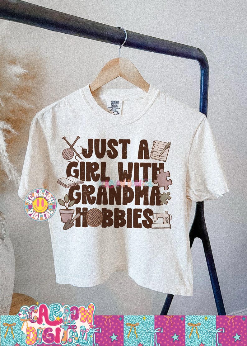 K&ouml;nnte beinhalten: Cremefarbenes T-Shirt mit dem Text "JUST A GIRL WITH GRANDMA HOBBIES" in Braun und bunten Buchstaben. Das Design zeigt Illustrationen von Stricknadeln, Notizbuch, Buch, Puzzleteil, Topfpflanze, N&auml;hmaschine und Garnkn&auml;uel, mit metrischen Ma&szlig;en.