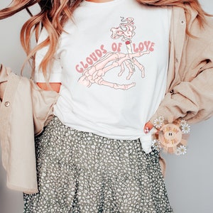 May include: White t-shirt with a graphic of a skeleton hand holding a lit cigarette, smoke forming hearts, and the text "Clouds of Love" in pink. The model is wearing a floral skirt and a tan corduroy jacket.