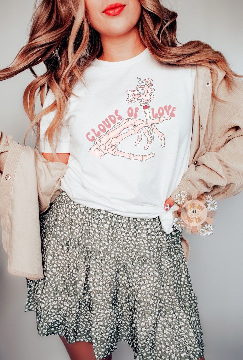 May include: White t-shirt with a graphic of a skeleton hand holding a lit cigarette, smoke forming hearts, and the text "Clouds of Love" in pink. The model is wearing a floral skirt and a tan corduroy jacket.