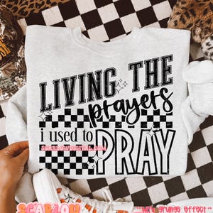 May include: A light grey sweatshirt featuring the text "LIVING THE prayers I used to PRAY" in black. The design incorporates a checkered pattern and star accents. The bottom of the sweatshirt is partially visible.