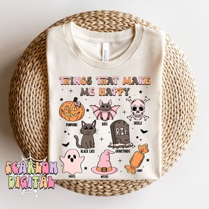 Puede incluir: Una camiseta blanca con un dise&ntilde;o gr&aacute;fico de tem&aacute;tica de Halloween. El texto "Things that make me happy" est&aacute; en la parte superior del dise&ntilde;o. El gr&aacute;fico incluye calabazas, murci&eacute;lagos, calaveras, un cementerio, gatos negros, fantasmas, brujas y caramelos.