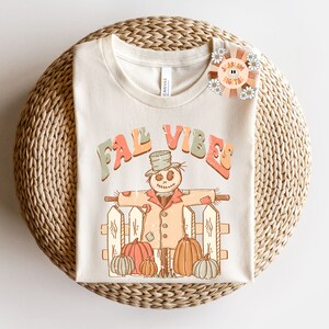 Op de afbeelding: Een beige T-shirt met de tekst "Fall Vibes" in kleurrijke letters. Een cartoon-vogelverschrikker met een hoed en een jas staat voor een hek met pompoenen.