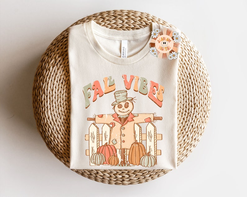 Op de afbeelding: Een beige T-shirt met de tekst "Fall Vibes" in kleurrijke letters. Een cartoon-vogelverschrikker met een hoed en een jas staat voor een hek met pompoenen.