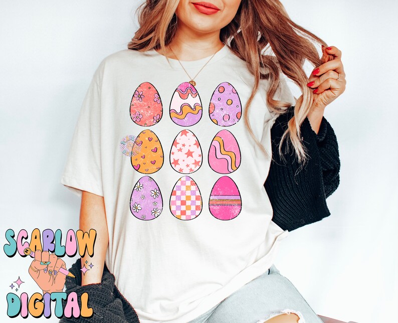 May include: A white t-shirt with a design of nine pastel Easter eggs with different patterns. The eggs are arranged in a 3x3 grid.