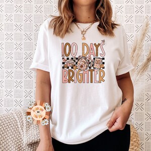 May include: White t-shirt with a graphic design that says "100 Days Brighter" in pink and gold lettering. The graphic also includes a checkered pattern and a floral design.