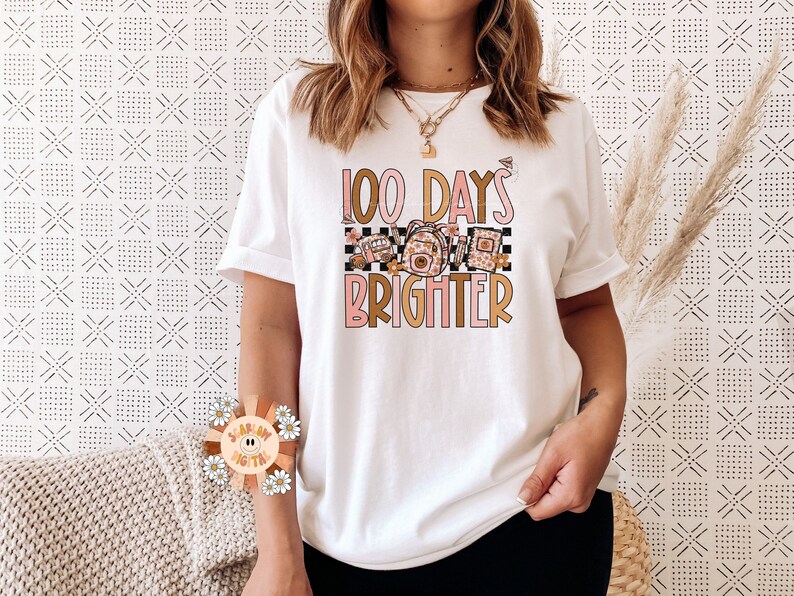 May include: White t-shirt with a graphic design that says "100 Days Brighter" in pink and gold lettering. The graphic also includes a checkered pattern and a floral design.