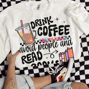 May include: White sweatshirt with the text "DRINK COFFEE avoid people and READ BOOKS." The graphic includes a coffee cup with flowers and books. The sweatshirt is on a black and white checkered surface.