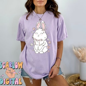 May include: A lavender t-shirt with a white bunny wearing a pink bow and surrounded by pink flowers. The bunny is smiling and has a cute expression.
