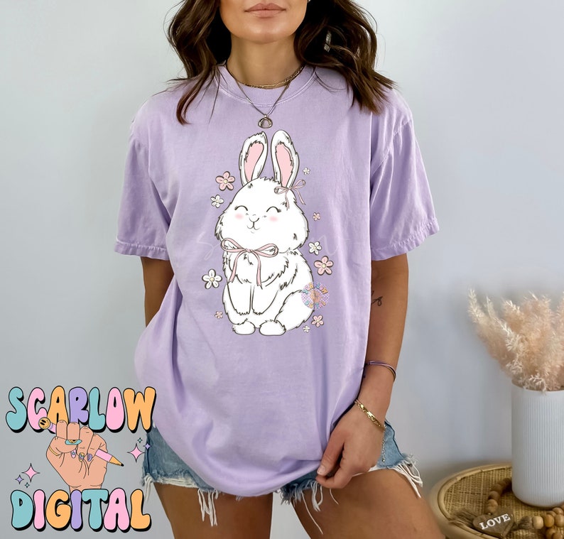 May include: A lavender t-shirt with a white bunny wearing a pink bow and surrounded by pink flowers. The bunny is smiling and has a cute expression.