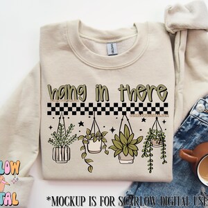 K&ouml;nnte beinhalten: Beiges Sweatshirt mit dem Text "hang in there" in Gr&uuml;n und Schwarz. Darunter ein schwarz-wei&szlig;es Schachbrettmuster und Illustrationen von H&auml;ngepflanzen. Das Sweatshirt wird mit einer Jeans und einer Kaffeetasse gezeigt.