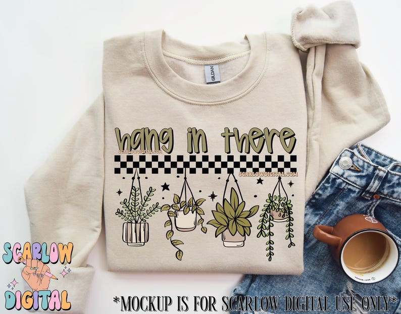K&ouml;nnte beinhalten: Beiges Sweatshirt mit dem Text "hang in there" in Gr&uuml;n und Schwarz. Darunter ein schwarz-wei&szlig;es Schachbrettmuster und Illustrationen von H&auml;ngepflanzen. Das Sweatshirt wird mit einer Jeans und einer Kaffeetasse gezeigt.