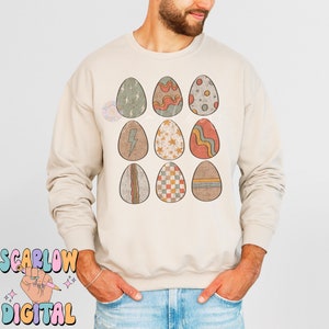 May include: A beige sweatshirt with a graphic of nine colorful Easter eggs with different patterns. The eggs are arranged in three rows of three.