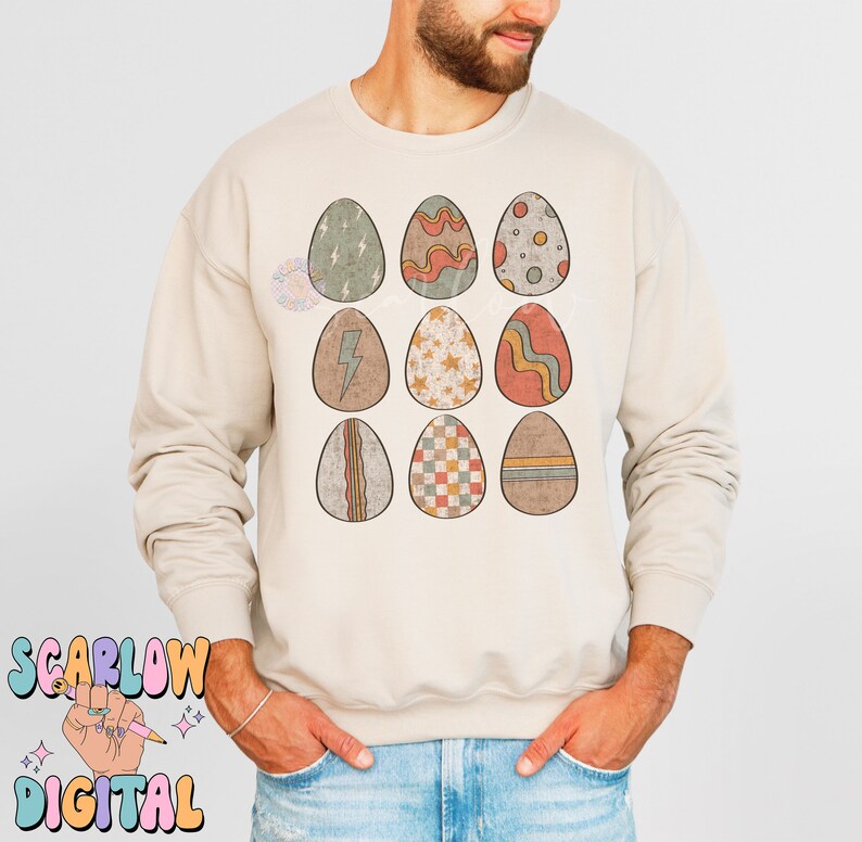 May include: A beige sweatshirt with a graphic of nine colorful Easter eggs with different patterns. The eggs are arranged in three rows of three.