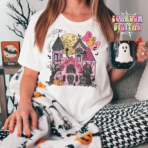 May include: A white t-shirt with a pink and black illustration of a haunted house with a crescent moon and bats. The house has a cross on the door and a pumpkin in front of it.