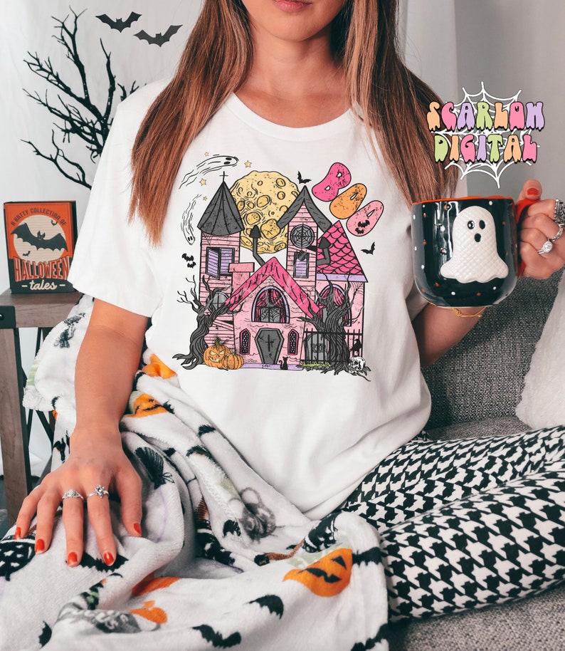 May include: A white t-shirt with a pink and black illustration of a haunted house with a crescent moon and bats. The house has a cross on the door and a pumpkin in front of it.