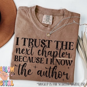 May include: A brown t-shirt with the text "I trust the next chapter because I know the author" printed in black. The shirt is on a table with a brown hat and a gold necklace.