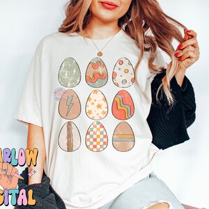May include: A white t-shirt with a graphic of nine colorful Easter eggs with different patterns. The eggs are arranged in a 3x3 grid.