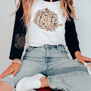 Pu&ograve; includere: Una maglietta da baseball bianca e nera con un design a forma di zucca. La zucca &egrave; decorata con fiori e il testo "Pumpkin Season" &egrave; scritto in un font retr&ograve;.